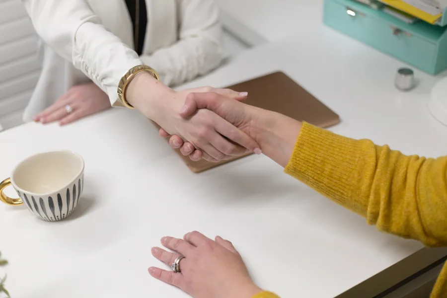 Two people shaking hands across a desk beside a cup of coffee